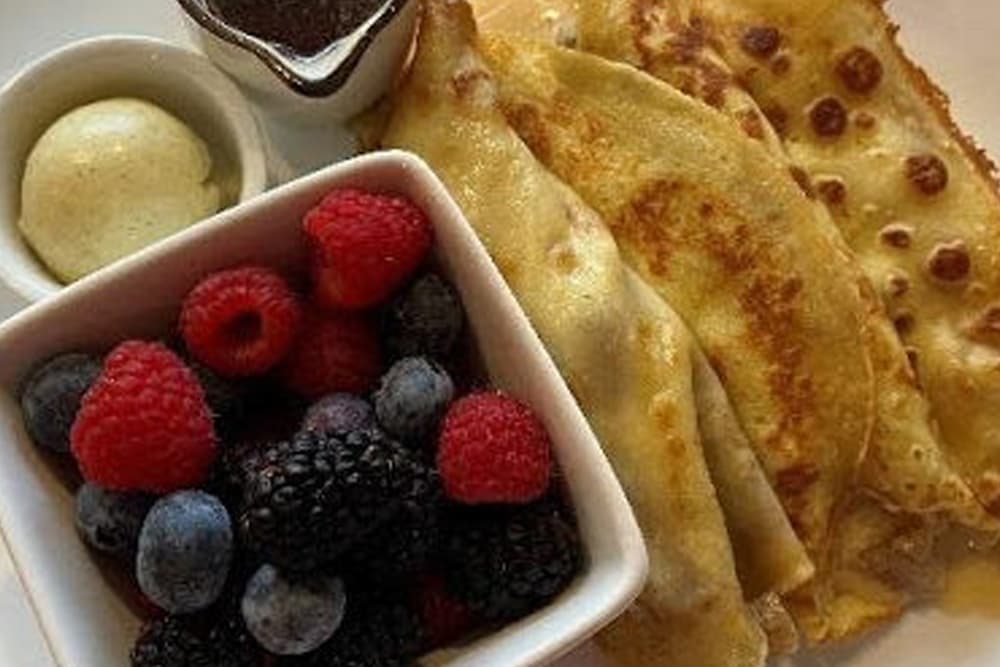 A delicious breakfast plate with crepes served alongside fresh berries, butter, and a small dish of jam. The vibrant colors of raspberries, blueberries, and blackberries enhance the simple, elegant meal.