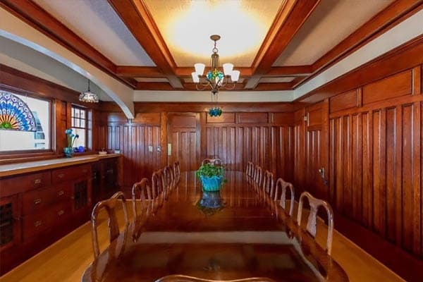 A warm dining room with wood paneling, beamed ceilings, a polished table, elegant chairs, chandelier, and stained glass window.