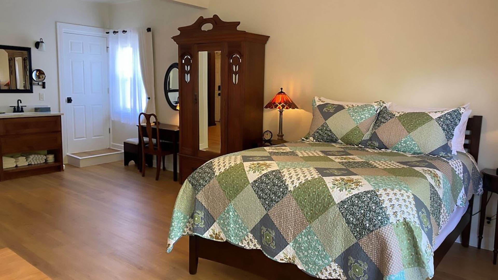 A cozy bedroom features a large wooden bed with a patchwork quilt. An antique wardrobe, vintage desk, and lamp add a rustic touch. Natural light brightens the simple, inviting space.